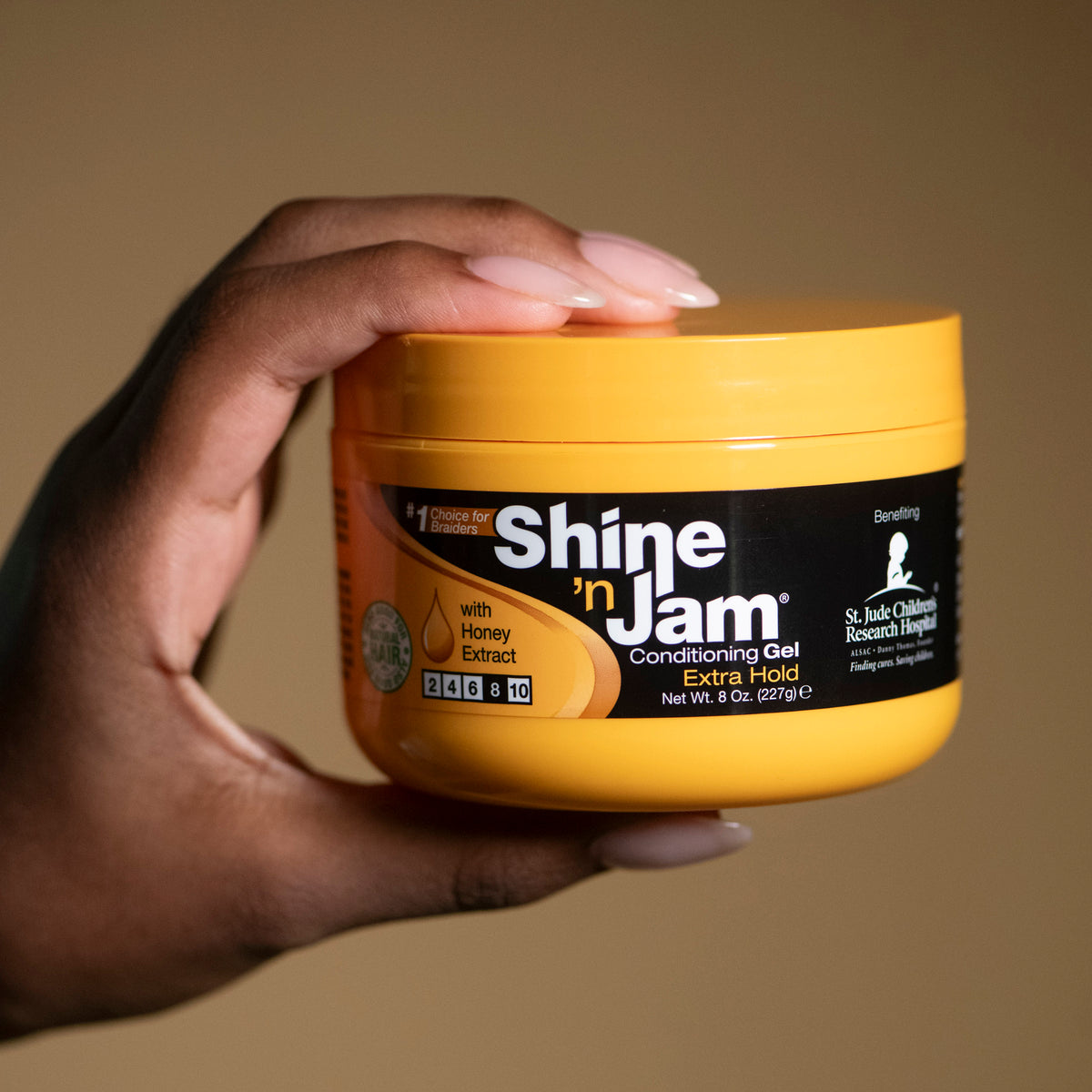Eine Hand hält ein gelbes Glas Ampro Shine 'n Jam Conditioning Gel. Auf dem Glas ist eine schwarze Beschriftung und das Bild einer Honigwabe zu sehen. Das Produkt soll dem Haar zusätzlichen Halt und Glanz verleihen.