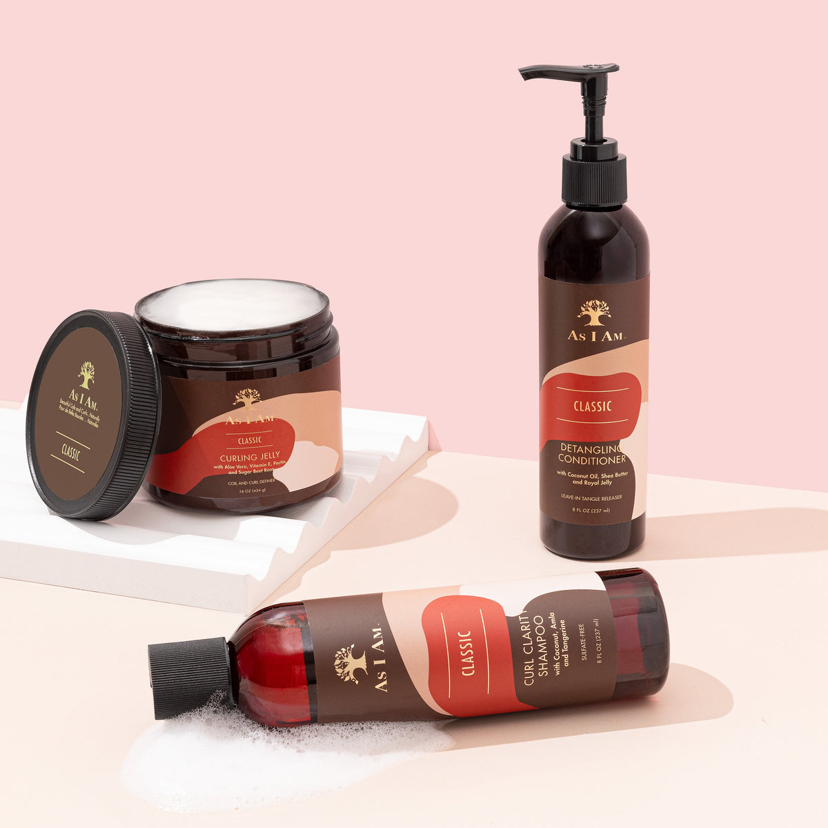 Drei Haarpflegeprodukte von As I Am stehen auf einer hellen Tischplatte. Von links nach rechts sieht man eine kleinere Flasche Shampoo, ein großes Glas Haarcreme und eine große Flasche Spülung. Die Produkte haben alle eine braune Farbe und ein weißes Etikett mit goldenen Details.