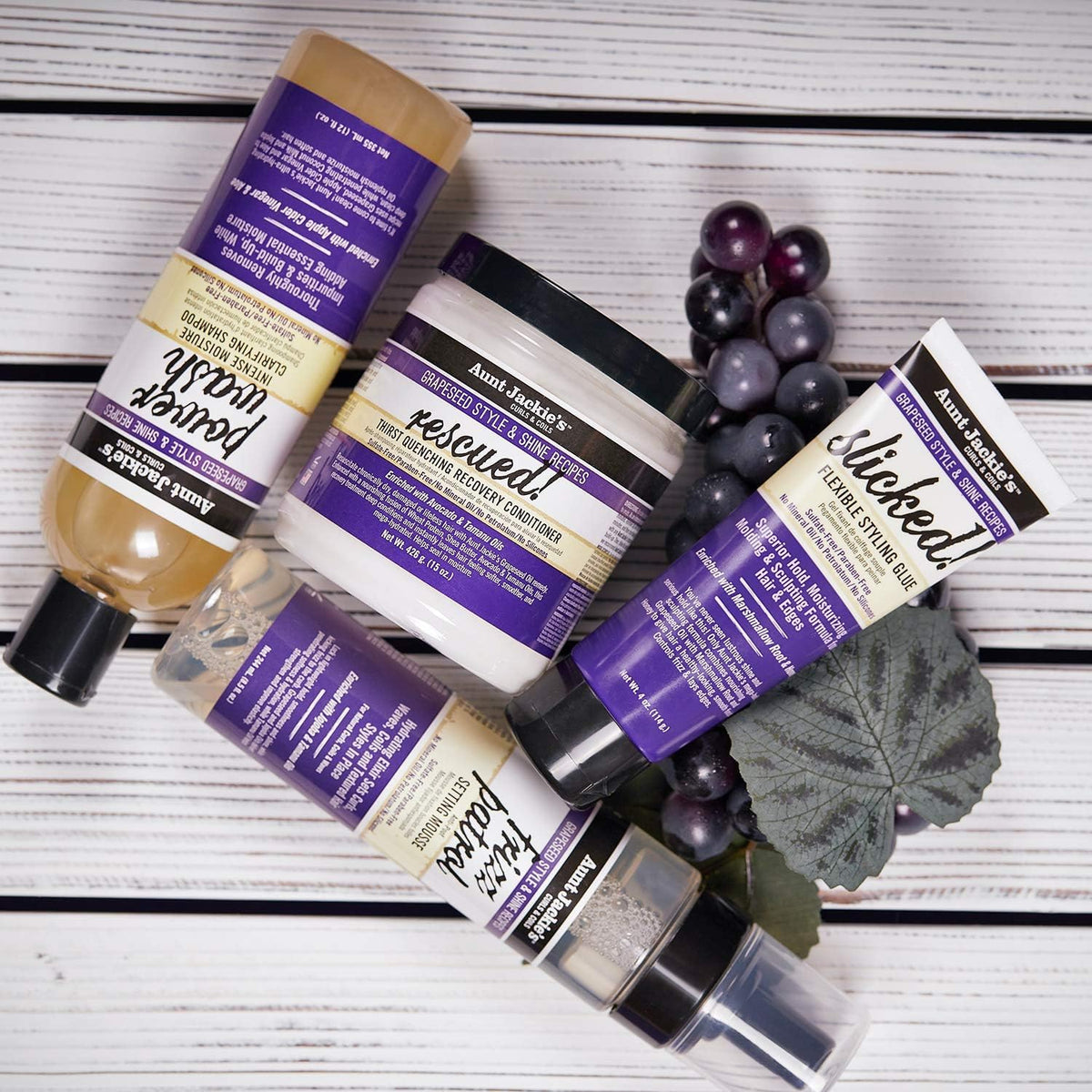Auswahl an Haarprodukten von Tante Jackie: Power Wash Shampoo, Rescued Conditioner, Slicked Styling Glue und Frizz Patrol Mousse, begleitet von dunklen Trauben.