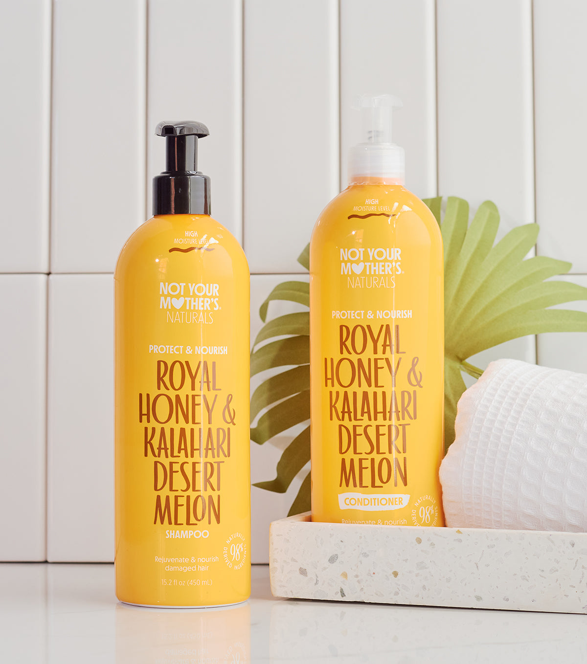 Zwei gelbe Flaschen: Not Your Mother's Naturals Royal Honey & Kalahari Desert Melon Shampoo (links) und Spülung (rechts), stehend auf einer weißen Fläche vor einer weißen Fliesenwand.
