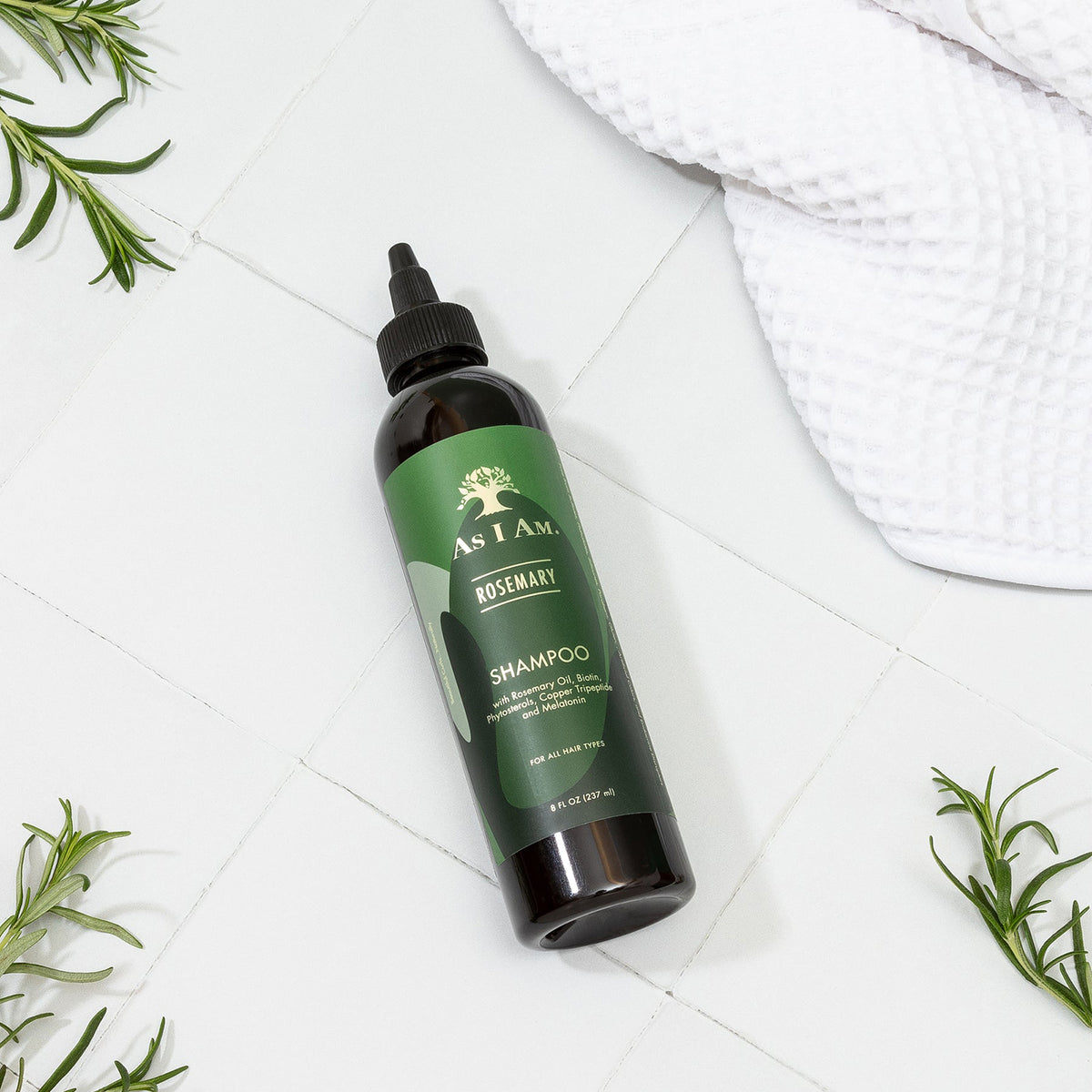 Eine Flasche As I Am Rosemary Shampoo (237 ml) liegt schräg auf einer weiß gefliesten Oberfläche, umgeben von frischen Rosmarinzweigen und einem weißen Handtuch.