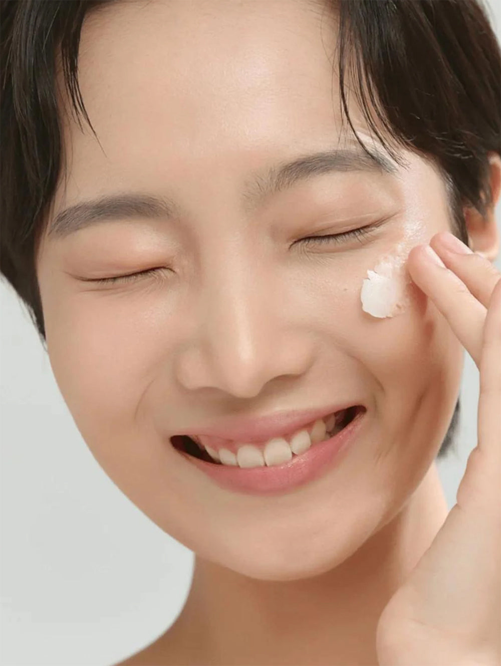 Lächelnde Frau mit geschlossenen Augen trägt Beauty of Joseon Radiance Cleansing Balm 100 ml mit der Fingerspitze auf die Wange auf.