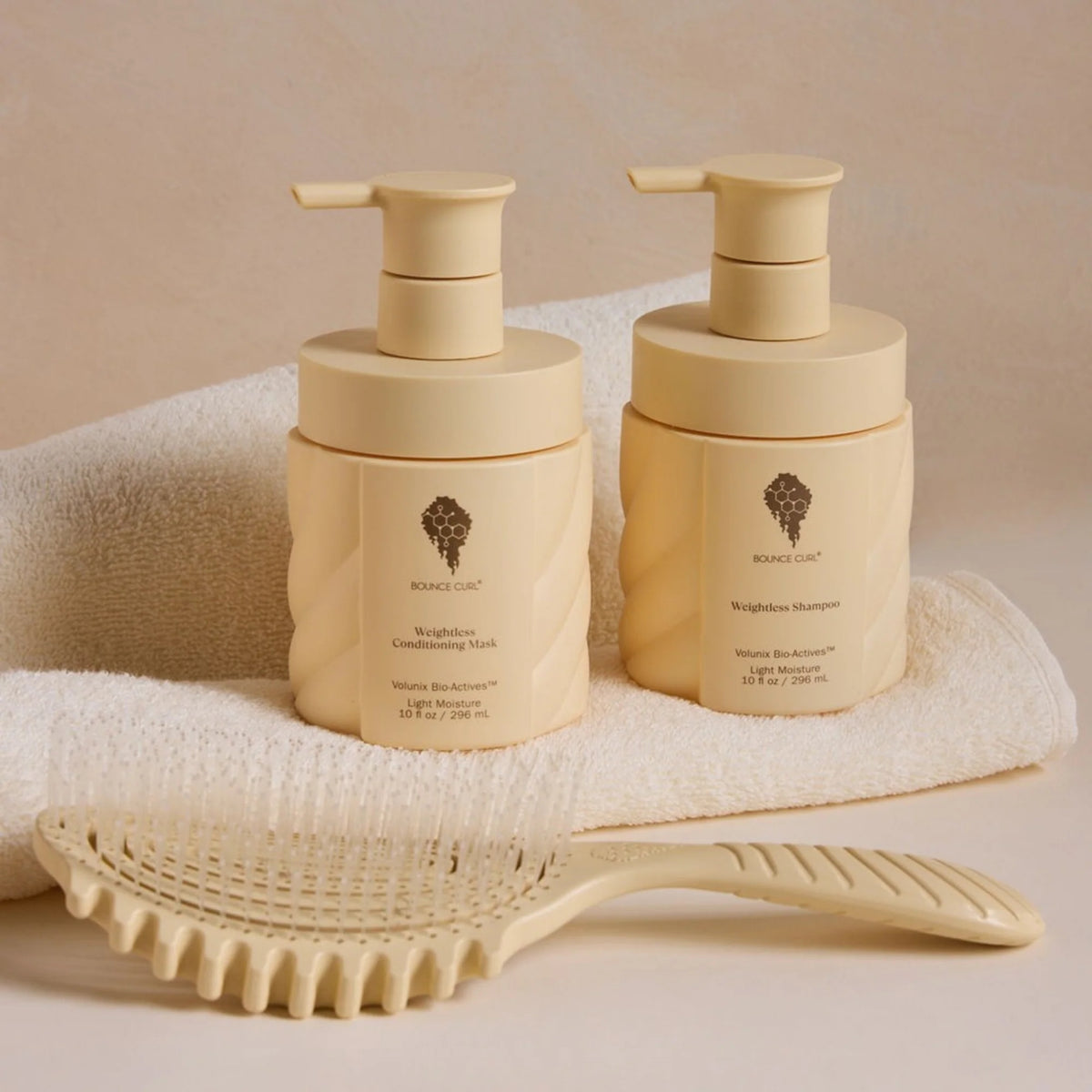 Bounce Curl Weightless Shampoo und Conditioning Mask in Cremeflaschen mit Pumpe, auf ein weißes Handtuch neben der Haarbürste legen.