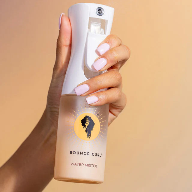 Handgehaltener Bounce Curl Water Mister mit weißer Sprühdüse und Logo vor beigem Hintergrund.