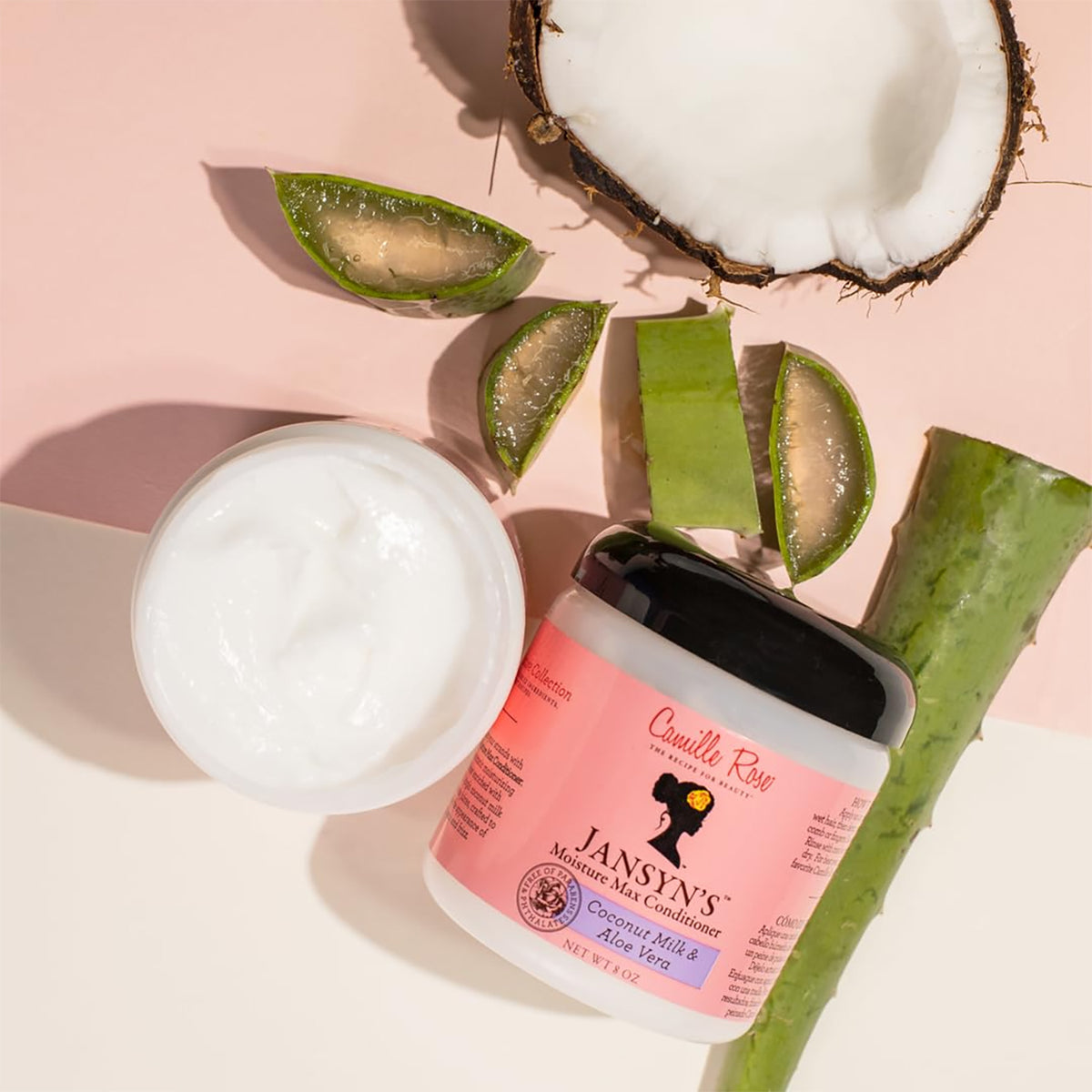 Camille Rose Jansyns Moisture Max Conditioner in einem weißen Behälter mit schwarzem Deckel. Das Produkt wird zusammen mit natürlichen Inhaltsstoffen wie Kokosnuss und Aloe Vera gezeigt.