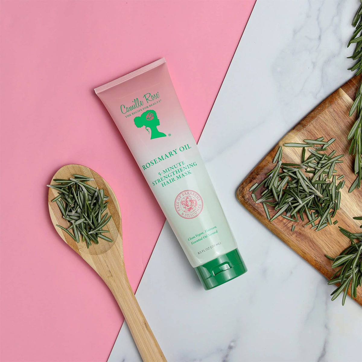 Eine Tube Camille Rose Rosemary Oil 5-Minute Strengthening Hair Mask liegt auf einem rosa Hintergrund neben einem Holzlöffel voller frischer Rosmarinblätter und einem Schneidebrett mit zusätzlichem Rosmarin.