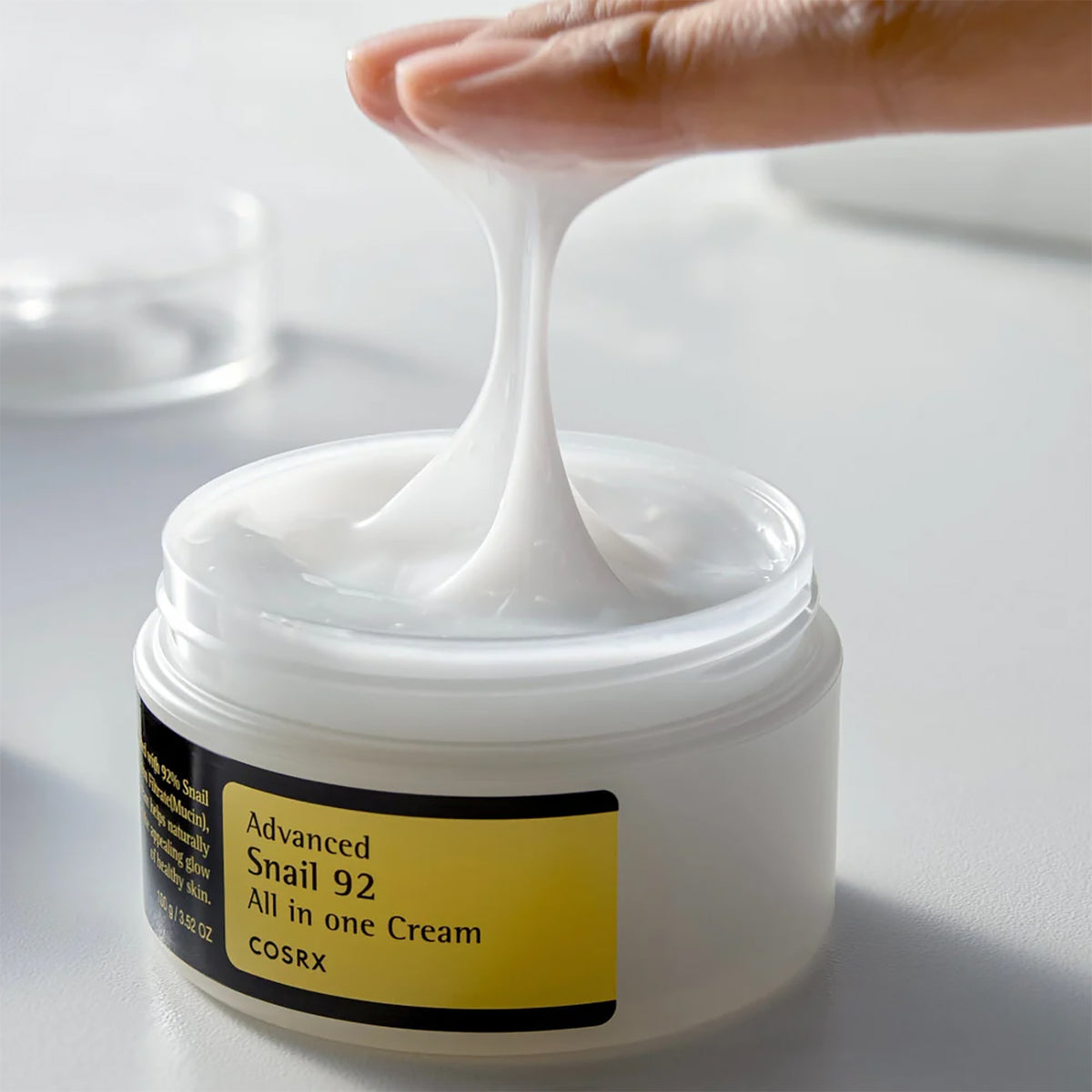 Finger heben elastische Textur aus offenem Glas Cosrx Advanced Snail 92 Cream 100gr auf weißem Tisch.