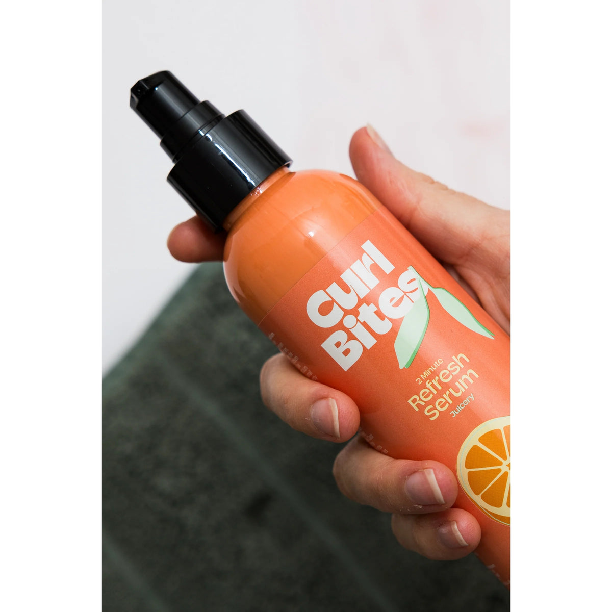 Hand hält orangefarbene Curl Bites 2 Minute Refresh Serumflasche mit schwarzer Pumpe vor grünem Hintergrund.