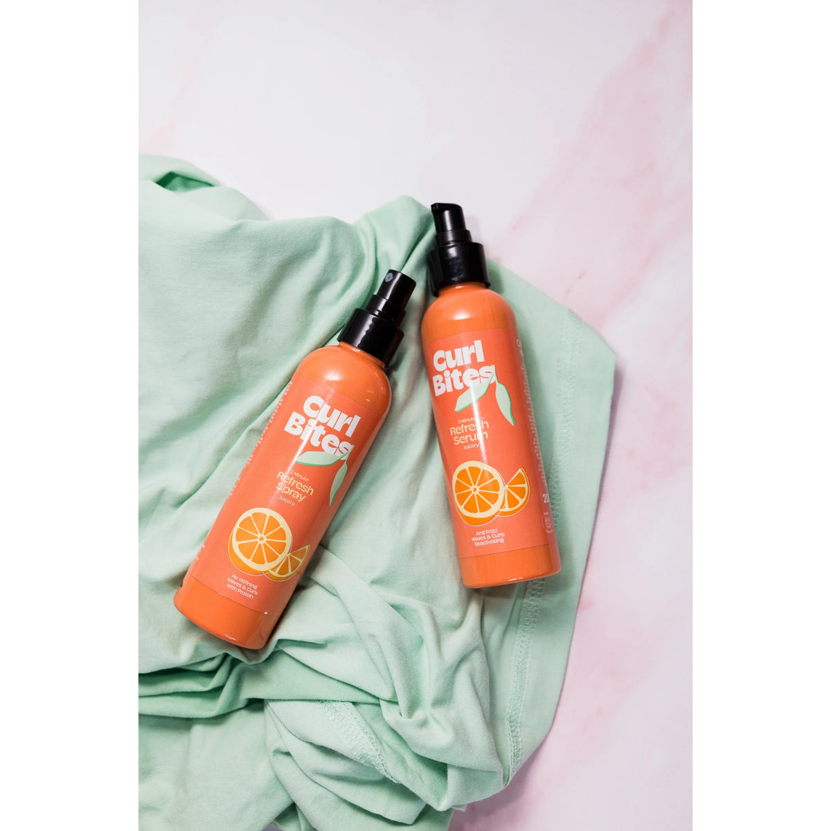 Curl Bites Refresh Spray und Refresh Serum in orangefarbenen Flaschen mit schwarzen Pumpverschlüssen, platziert auf einem mintgrünen Stoffhintergrund.