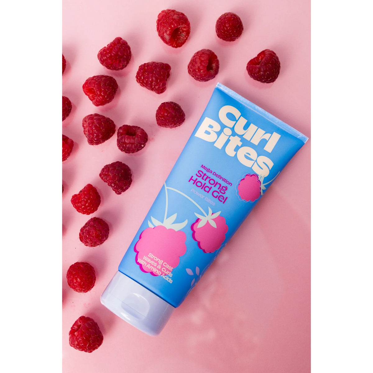 Curl Bites Strong Hold Gel in blauer Tube mit Himbeeren, umgeben von frischen Himbeeren auf rosa Hintergrund.