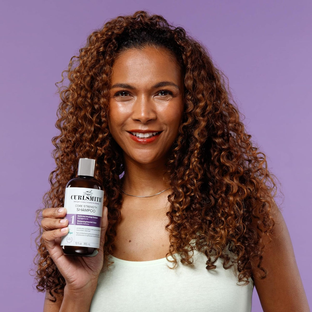 Frau mit lockigem Haar lächelt und hält Curlsmith Core Strength Shampoo vor violettem Hintergrund.