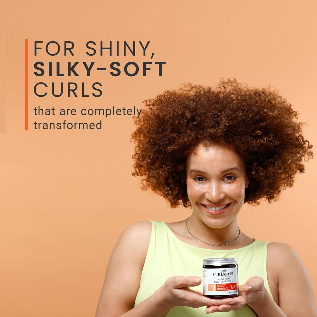 Eine Frau mit kastanienbraunen Locken lächelt und hält den Curlsmith Double Cream Deep Quencher für glänzende, seidige Locken.