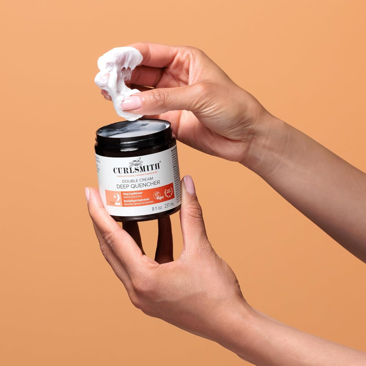 Die Hand nimmt die cremige Curlsmith Double Cream Deep Quencher Haarkur aus einem schwarzen Glas vor orangefarbenem Hintergrund auf.