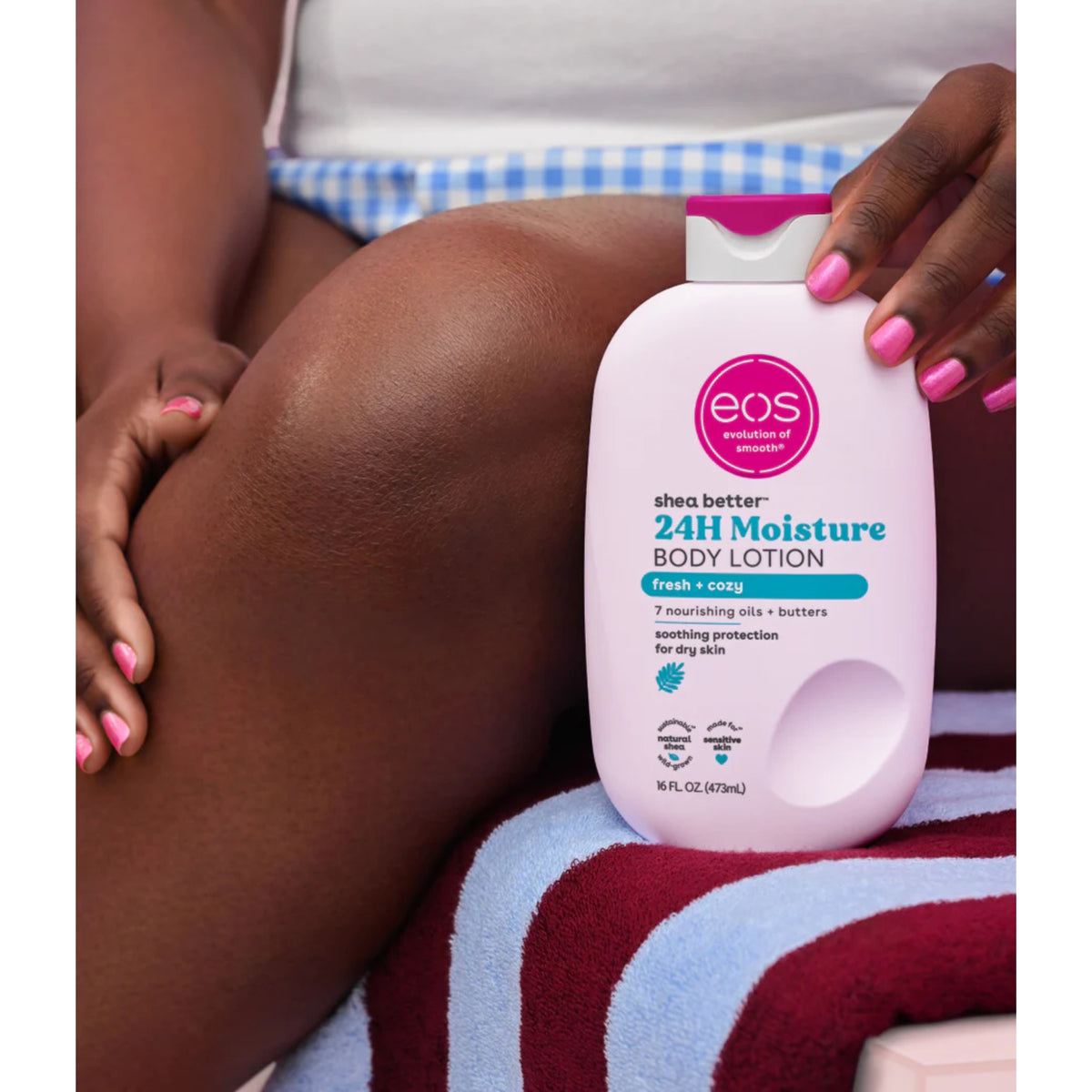 eos Shea Better 24H Moisture Body Lotion in einer weißen Flasche mit rosa Verschluss, die auf einem gestreiften Handtuch an das gebräunte Bein gehalten wird.