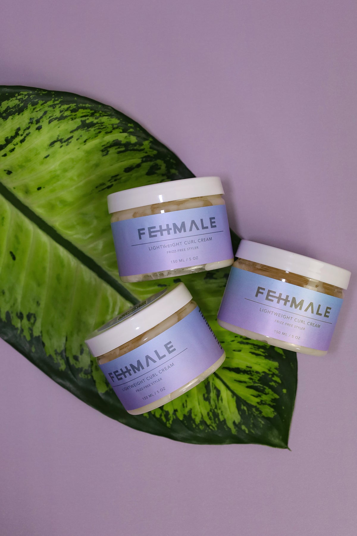 Drei Fehmale Lightweight Curl Cream 150-ml-Gläser auf grünem Blattdekor vor violettem Hintergrund, perfekt für lockiges Haar.