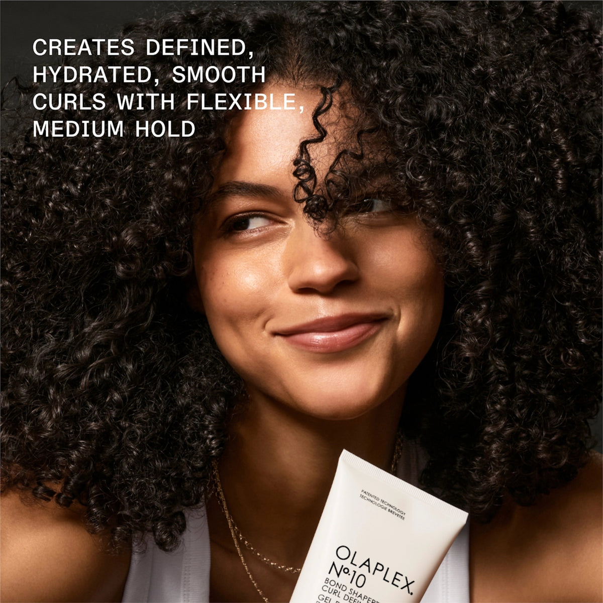 Eine lächelnde Frau mit definierten Locken hält das Olaplex No.10 Bond Shaper Curl Defining Gel für hydratisierte Locken mit flexiblem Halt.