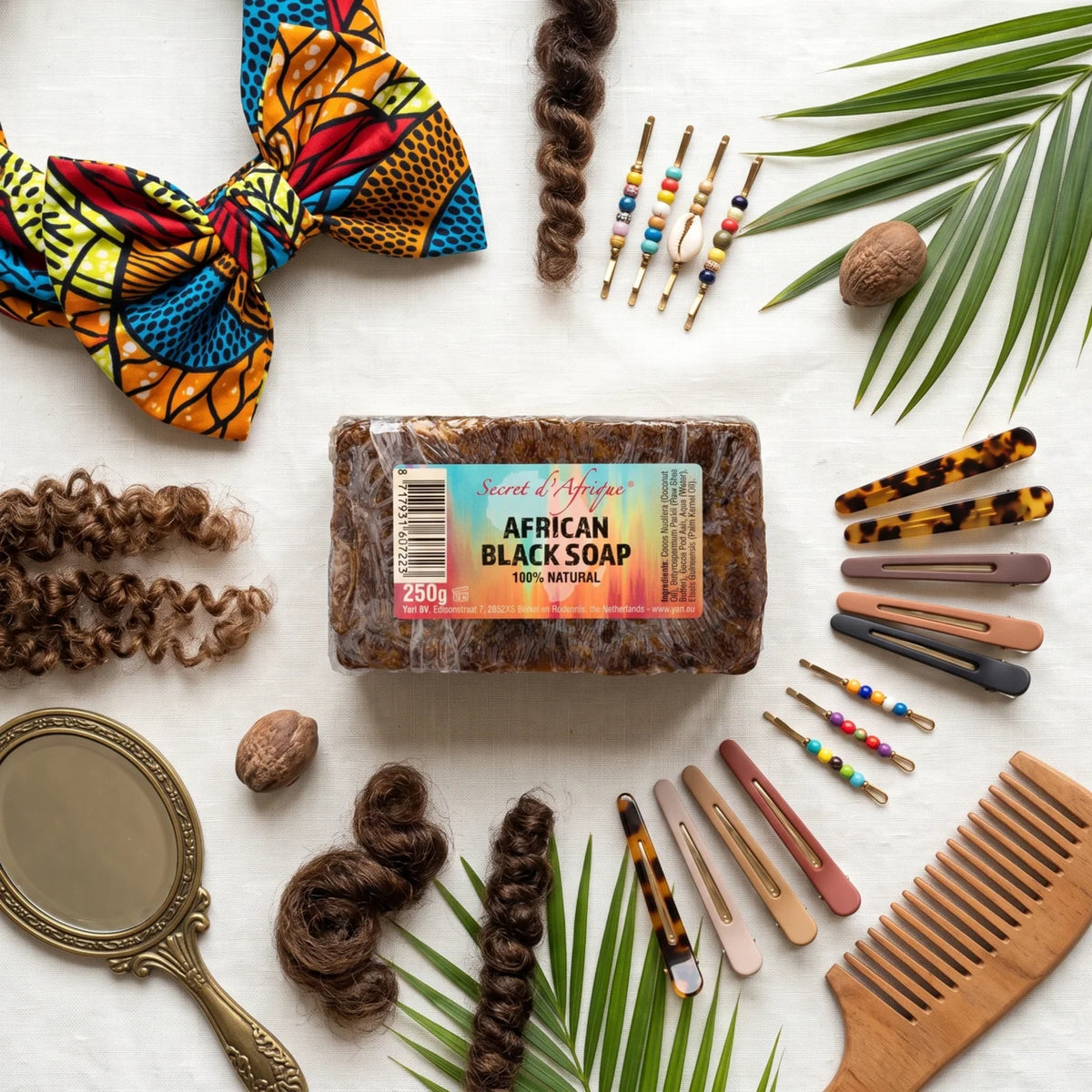 Secret d'Afrique Afrikanische schwarze Seife 250 g, umgeben von Haarspangen, afrikanischem Turban, lockigem Haar und hölzernem Haarkamm.