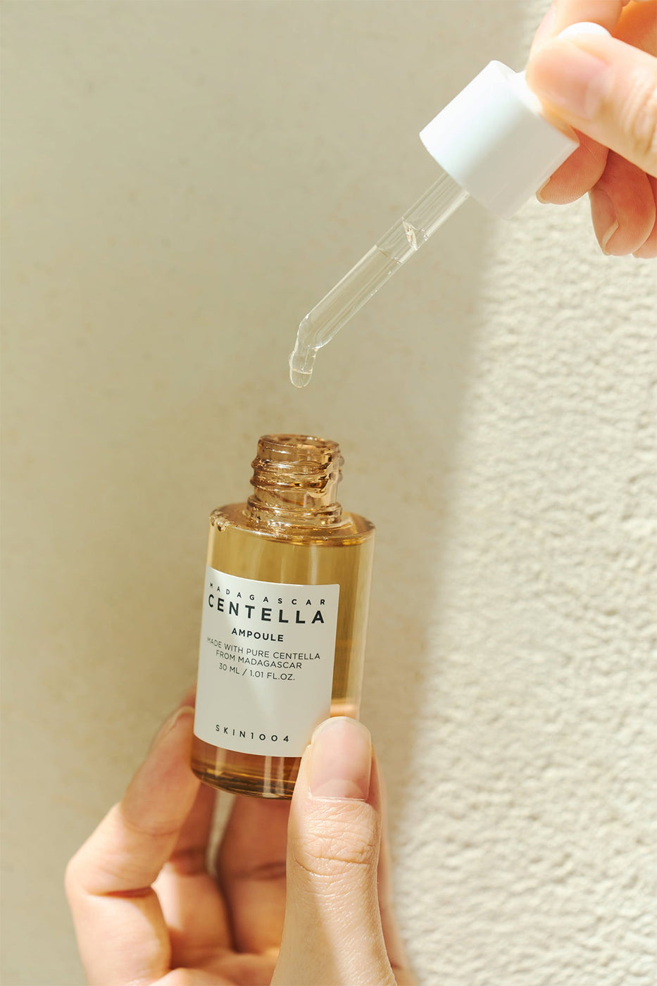 Pipette mit Serum von SKIN1004 Madagascar Centella Ampoule 100 ml über einer goldenen Flasche im Sonnenlicht gegen eine helle Wand gehoben.